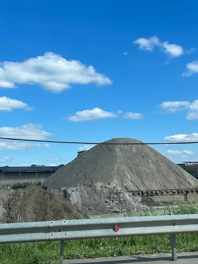 The image depicts an outdoor industrial or construction site under a bright blue sky with scattered white clouds. In the foreground, a metallic guardrail runs horizontally across the bottom of the frame, with green grass and some small white flowers growing behind it. Behind the guardrail, a large, conical pile of grey material, possibly sand or gravel, dominates the midground. To the left of this large pile is a smaller mound of darker, loamy soil. In the background, a long, low-rise building with a dark roof and lighter walls is visible, partially obscured by the piles of material. A dark, thick cable or pipe stretches horizontally across the upper portion of the image, in front of the sky and the building. The overall scene suggests a location involved in material storage or processing, possibly a quarry or a construction material depot. The specific location in the city of Верхнерусское, Russia, is not discernible from the visual elements alone.