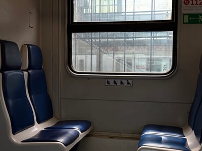 The image shows the interior of a train car, likely a commuter train, with rows of blue and white seats. Two seats are visible in the foreground on the left, and parts of two seats are visible on the right. The seats appear to be made of vinyl or a similar material. Through the window, other train cars and a station structure are visible. On the wall above the window, there are signs indicating "112 EMERGENCY SERVICE" and an arrow pointing left with a figure of a person. Below the window, there are small icons depicting people sitting. The environment is dimly lit, suggesting an overcast day or the interior lighting of the train. The location is identified as Aprelevka, Russia, indicating this is a train car operating in that region.