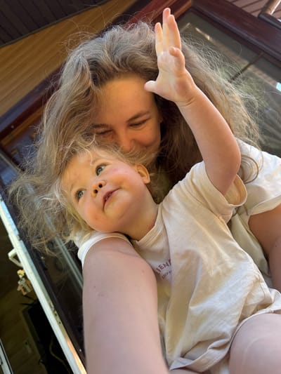 The image captures a close-up portrait of an adult female and a young child in what appears to be an outdoor or porch setting in Копнино, Russia. The adult, with long, wavy, light brown hair, is positioned behind and to the left of the child, with her face partially visible, angled downwards. Her eyes are visible in some crops, and she appears to be looking at the child. Her right arm is around the child. The child, a fair-skinned toddler with blonde, curly hair and blue eyes, is in the foreground, looking upwards and slightly to the right with a soft expression. The child's left hand is raised with fingers splayed, positioned near the adult's head. Both individuals are wearing light-colored, short-sleeved garments; the child's shirt has a faded, reddish-brown text partially visible on the left side, reading "EN" and "KEEP H". The background consists of dark brown wooden structures, likely a house exterior or a door/window frame, with a white door frame and a metallic handle/lock mechanism visible on the far left. The scene is illuminated by natural light.