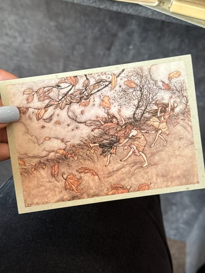 A postcard-sized illustration depicts four young females with long, flowing hair, engaged in a dynamic dance amidst falling autumn leaves and bare branches. The scene has a whimsical, ethereal quality, rendered in muted earth tones with delicate linework. The figures are barefoot and dressed in flowing gowns, their bodies contorted in movement as if caught by a gust of wind. The background is a muted wash of color with subtle shading, suggesting a natural, outdoor setting. The postcard is held by a hand with light grey painted fingernails, against a dark, textured background that appears to be fabric. There is no identifiable location within the city of Verkhnii Lars, Russia, or any other specific geographical context present in the image.