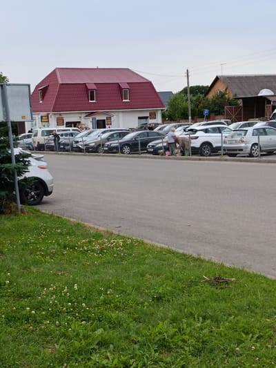 The image depicts an outdoor scene in a parking lot adjacent to a building with a distinctive red mansard roof. Several cars are parked in rows. In the mid-ground, two individuals are interacting near a white car and a light-colored sedan. One person appears to be bending down, possibly retrieving items from a bag, while the other stands nearby. A building with multiple windows and a prominent red roof is visible on the left side of the image, and other buildings and trees are present in the background. A parking sign with a 'P' symbol is visible on a pole. The foreground consists of a paved road and a grassy area with small white flowers. The sky is overcast. The scene is consistent with a commercial or residential area in Смілавіцкі сельскі Савет, Belarus.
