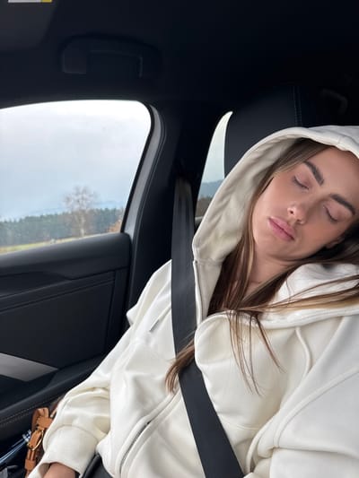 The image shows the interior of a vehicle, specifically the front passenger side, located in the city of Hetzendorf, Austria. A female subject is seated in the passenger seat, appearing to be asleep with her eyes closed and head tilted. She has long, light brown hair, partially visible from beneath the hood of an off-white, zippered sweatshirt she is wearing. A black seatbelt is fastened across her chest and lap. The vehicle's interior features dark-colored upholstery, and an overhead grab handle is visible above the window. Through the passenger window, an overcast sky is visible above a landscape of distant tree-covered hills or mountains, with a single leafless tree in the middle ground, indicating a rural or natural setting. A small, light brown object, possibly a bag, rests on the seat near the subject's lap.