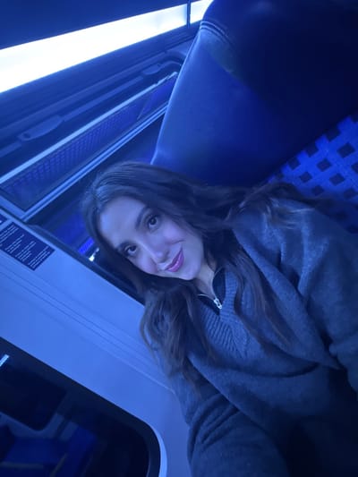 A young woman sits in a train car. She wears a gray zip-up sweater and has long brown hair. The train car has blue patterned seats and a large window with bright light shining through. The woman is smiling slightly and looking at the camera. The image has a blue tint to it.