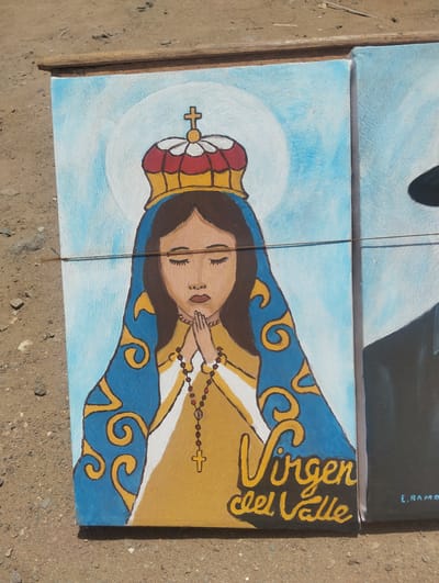 The image shows a painting of the Virgin del Valle, displayed outdoors. The painting depicts the Virgin Mary with a crown, blue and gold robe, and a rosary. The words "Virgen del Valle" are written in gold paint at the bottom right. The painting appears to be resting on the ground, next to another painting that is partially visible. The ground is made up of soil and small rocks. Given the name of the Virgin, it is likely located in El Dátil, Venezuela, where she is a patron saint.