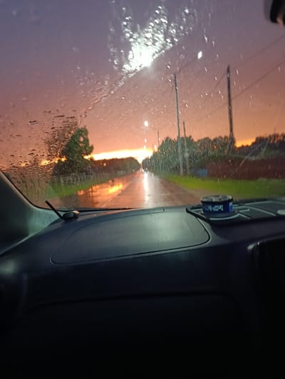 The image shows a view from inside a car, looking through a rain-streaked windshield at dusk. The sky is a gradient of purple and orange, with the sun setting on the horizon. The road ahead is wet, reflecting the colors of the sky. Trees and power lines are visible along the side of the road. On the dashboard, there is a small blue container with a colorful lid. The overall mood is one of a quiet, reflective drive during a rainy evening.