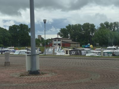 The image depicts a harbor in Šilutė, Lithuania, with several boats docked. The foreground shows a cobblestone walkway with a tall gray utility pole. In the background, a cluster of buildings, including what appears to be a boathouse or marina facility, is visible. Trees line the background, and the sky is overcast with dramatic clouds. The boats are a mix of recreational vessels, with several featuring white hulls and canvas coverings. The overall atmosphere is that of a typical harbor scene on a cloudy day.