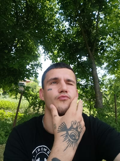 The image shows a close-up, self-taken photograph of a young man outdoors, likely in a rural or garden setting, suggested by the presence of lush green trees and foliage. The man is looking towards the camera with his right eye open and his left eye squinting. He has his left hand resting on his chin and cheek.
The man has a tattoo on his left cheek, which appears to be text. He is wearing a black t-shirt with a circular logo that has white text inside. On his right hand, he has a prominent tattoo of an eye with rays emanating from it, and a spiderweb design on his thumb. He is also wearing a silver chain necklace.
The lighting suggests it is daytime, with sunlight filtering through the leaves of the trees, creating a dappled effect. The overall mood appears relaxed and casual, with the man making a slightly playful expression. The background is filled with greenery and some fence posts, implying an outdoor location. Given the location context of Soroca, Moldova, the scene likely represents a warm, pleasant day.