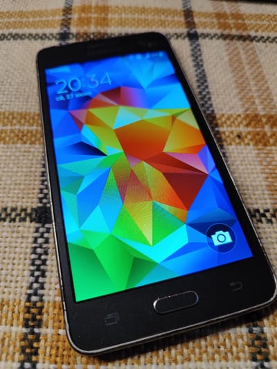A Samsung Galaxy S5 smartphone is displayed on a checkered fabric surface. The phone is in landscape orientation, with its screen turned on. The screen shows a colorful, abstract wallpaper with geometric patterns in shades of blue, green, orange, and red. The time displayed on the screen is 20:34, and the date is 06.27. A camera icon is visible on the bottom right of the screen. The phone has a black bezel and a silver trim. The fabric surface appears to be a tablecloth or placemat with a plaid pattern of brown, yellow, and black threads. The image was taken in Korolyov, Russia.