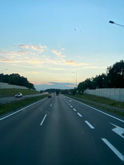 This image depicts a view from a vehicle traveling on a multi-lane highway, likely in or near Zakrzów, Poland, during twilight. The sky is a gradient of light blue, transitioning to a soft orange and pink hue near the horizon, with scattered clouds. Two small aircraft, possibly planes, are visible in the upper right portion of the sky. The highway has clear lane markings, with white dashed lines delineating the lanes and solid white lines along the edges. Several vehicles, including cars and a truck, are traveling on the highway. On either side of the road, there are sound barriers and dense green trees. Streetlights are intermittently placed along the highway. The overall impression is one of a peaceful, early evening drive on a well-maintained road, set against a scenic natural backdrop.