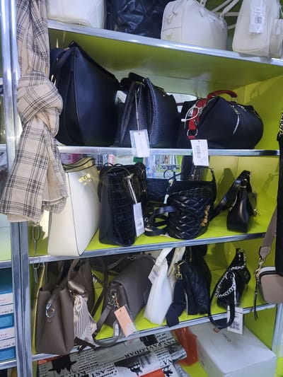 This is a medium-shot, eye-level, interior photograph of a retail display of handbags. The handbags are arranged on bright yellow shelves that are divided by metal poles. The shelves are well-lit, and the focus is on the merchandise.
On the top shelf, there are three handbags. On the left is a large black handbag with a textured surface. Next to it is a black handbag with a woven pattern and a red handle. On the right is a black handbag with a red handle and zipper detail.
The middle shelf has several handbags. On the left is a large white handbag. To its right is a black handbag with a crocodile-like texture. Next to that is a black handbag with a quilted pattern and a gold logo. Further to the right is a black shoulder bag.
The bottom shelf displays several more handbags. On the left is a brown handbag with a patterned scarf tied to the handle. Next to it is a gray handbag with a black strap. To the right are several other handbags, some with chain straps.
In the background, partially visible, are stacks of boxes, some with text and logos. The overall impression is a neat and organized retail environment focused on showcasing a variety of handbags. The location is identified as Ushtobe, Kazakhstan.