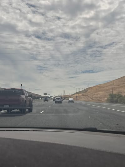 A daytime, eye-level, medium shot captures a view from the driver's perspective on a multi-lane highway. The road ahead is paved, with white dashed lines marking the lanes. Several vehicles are visible on the highway, including a red pickup truck in the left lane, a white car in the right lane further ahead, and other vehicles in the distance. The landscape features rolling hills on the right side, covered in dry grass, and a cloudy sky with scattered blue patches overhead. Electrical poles and wires are present along the highway and on the hills. The foreground shows the dashboard of the vehicle from which the photo is taken. The scene is set in Fairfield, California, United States of America.
