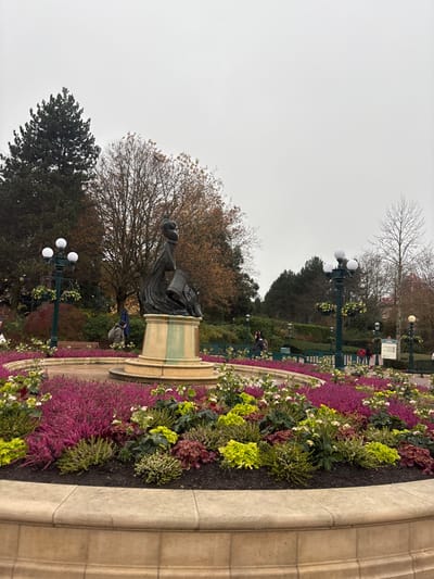 The image depicts a meticulously maintained outdoor park scene in Chessy, France, under an overcast, light gray sky. A large, circular, tiered flower bed, approximately 12 meters in diameter, dominates the foreground and mid-ground. This flower bed is bordered by a light beige stone curb and is densely planted with various ground covers and shrubs, predominantly featuring vibrant magenta-purple blooms, lime green foliage, and some darker green and reddish-brown plants.
At the precise center of this circular arrangement stands a dark-colored, likely bronze, sculptural fountain. The sculpture features a stylized vertical form, suggestive of a figure, with a distinct curving element at its apex and a cascading basin feature below. This sculpture is mounted on a multi-tiered, light beige stone pedestal, the lower sections of which display noticeable green discoloration, consistent with mineral deposits or algae from water exposure.
Around the perimeter of the central flower bed, several dark green lampposts with multiple spherical white globes are positioned, some featuring hanging flower baskets. Paved pathways, light gray in color, are visible in the mid-ground, encircling the flower bed and extending into the background.
Several individuals are present in the scene. One person, wearing a blue jacket and dark backpack, stands behind the fountain to the left. Multiple other figures are dispersed across the pathways in the mid-ground and background, appearing to be walking or standing. Their specific actions and interactions are not discernible.
The background features a diverse array of trees: tall, dark green coniferous trees are prominent on the left, while deciduous trees with sparse, reddish-brown and orange foliage, consistent with late autumn or early winter, are distributed across the center and right. Some completely bare deciduous trees are also visible on the far right. Dark green park benches are situated along the pathways in the background. A white information sign is partially visible on the far right, near a distant figure.