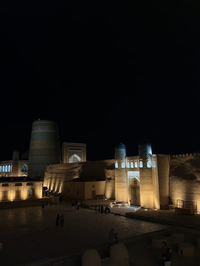 The image shows the Kalta Minor Minaret and the West Gate of the Ichon-Qala, the walled inner city of Khiva, Uzbekistan, illuminated at night. The minaret, a cylindrical structure with intricate blue and yellow tilework, stands prominently on the left. To its right is the imposing West Gate, a fortified structure with two domes topped with patterned tiles and arched windows that are lit from within. The outer walls of the Ichon-Qala, constructed of mud brick, are also illuminated by spotlights, casting long shadows and highlighting their texture. In the foreground, a courtyard area is visible, with several people walking and standing, some appearing to be tourists. The overall atmosphere is one of historical grandeur and a tranquil evening.