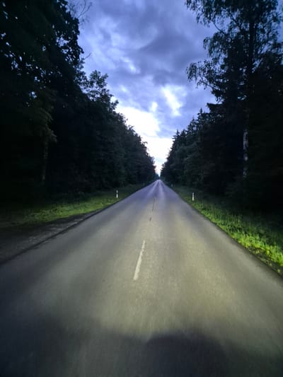 An image captures a straight asphalt road lined with dense trees, disappearing into the distance under a twilight sky. The road has a white dashed line down the center and is illuminated by headlights, casting a bright glow on the pavement. White reflective markers are visible on the sides of the road. The trees on either side are dark and create a tunnel effect, with the sky showing a mix of dark clouds and some lighter patches. The scene appears to be in a rural or wooded area. The location in the city of Krailling, Germany is not discernible from the image itself. There are no people, animals, or structures present, nor any discernable action or interaction. The environment is solely characterized by the road, trees, and sky.