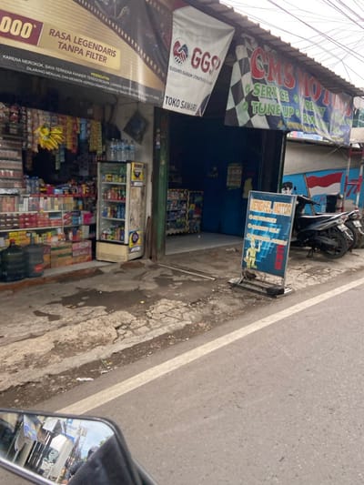 The image shows a street scene in Kecamatan Margahayu, Indonesia. On the left side of the street is a small shop with a sign reading "RASA LEGENDARIS TANPA FILTER" above the entrance. The shop has shelves of goods including cigarettes, snacks and drinks. There is a large refrigerator visible in the window. Across the street is a motorcycle repair shop with a sign reading "CMS MOTOR". The sign also states that they offer services including "SERVICE", "TUNE UP" and "REPAIR". There is a blue sign next to the shop with the words "BENGKEL MOTOR". A motorcycle is parked in front of the shop. There is a white line separating the road and the sidewalk. The sidewalk is dirty with debris and patches of grass.