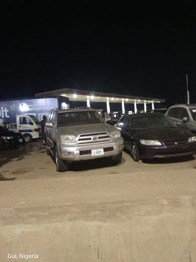 A night time, exterior, medium shot shows a row of parked vehicles at a Total Energies gas station. In the foreground, a silver Toyota 4Runner is parked to the left of a dark purple Honda Accord. The 4Runner has a Nigerian license plate that reads "RSH 62RR". To the left of the 4Runner, a white Toyota truck is parked in front of a large, dark truck with "Salt" and "Withdraw Here" printed on its side. Behind the parked vehicles, the gas station's canopy is illuminated by overhead lights. The ground is wet, indicating recent rain. In the bottom left corner of the image, "Gui, Nigeria" is written.