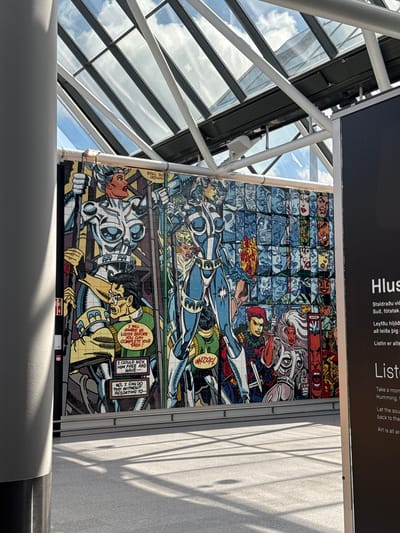 The image depicts an indoor public space, specifically the departure hall of an airport in Unknown, Iceland. A large, vibrant, comic book-style mural dominates the left side, featuring characters in futuristic armor with visible speech bubbles, against a backdrop of numerous stylized faces arranged in a grid. The mural displays text such as "STILL GOES DOWN," "I WILL SHAKE US LOOSE BEFORE YOU CAN COMPLETE YOUR TASK!," "I COULD KICK HIM FREE AND HAVE--," "NO. I CAN DO THIS WITHOUT RESORTING TO--," and "WHOOFL." Above the mural, a prominent glass ceiling allows natural light to enter, revealing a blue sky with white clouds through a grid of structural beams. A large, light grey cylindrical pillar stands on the far left, partially obscuring the mural and casting a distinct shadow on the light grey floor. On the far right, a dark informational stand is partially visible, displaying text in both Icelandic and English, with phrases like "Hlus...", "Staldraðu við...", "Listen...", and "Art is all ar...". Sunlight creates strong highlights and shadows across the scene.