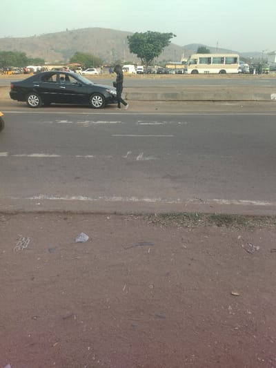A black car is parked on the side of a paved road. A man is walking away from the car, towards the right side of the image. The road is bordered by a concrete curb and a dirt shoulder. The road appears to be in a suburban or rural area, with hills in the background. There are other cars in the background, including a white bus. The image was taken at the coordinates 9.024, 7.573, which is located in the city of Maraba, Nigeria.  There is some debris and trash on the shoulder of the road.  The weather is cloudy and overcast.  The image appears to have been taken in the early morning or late afternoon, as the shadows are long.