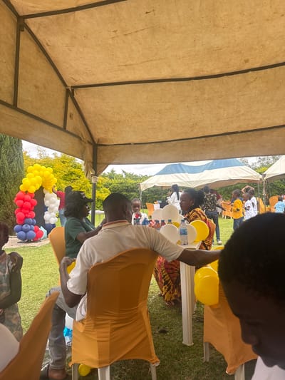 This media file shows an outdoor gathering under a tent, likely a celebration or community event. Several people are visible, mostly adults and some children, sitting and interacting. They are dressed in casual and festive attire, including traditional African fabrics and modern clothing. A table is set up with water bottles and balloons in yellow, white, red, and blue, suggesting a celebratory occasion like a birthday or party.
The scene is set on a grassy area, with trees and foliage visible in the background, indicating an outdoor location. The presence of a tent and the lighting suggest it is daytime, possibly late morning or afternoon. The mood appears relaxed and social, with people engaged in conversation and observing the event. Some individuals are wearing head coverings, and others have their hair styled in braids.
The overall impression is of a vibrant and joyful community gathering. The provided location context of Barikin Gidar Kanat, Nigeria, further helps to understand the cultural setting and the type of event that might be taking place. No specific text is clearly legible in the image.