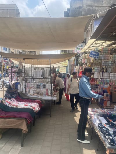 The image depicts an outdoor market scene in Ahmedabad, India, likely a busy street market. In the foreground, a stall displays folded textiles, including patterned fabrics in reds, blues, and greens. To the right, another stall is laden with various items, including packaged socks and what appear to be mobile phone cases, displayed on shelves and tables. A man in a blue denim shirt and black pants, wearing white shoes, is standing near this stall, reaching out his right hand towards the merchandise. Further into the market, a man in a light-colored t-shirt and glasses walks away from the camera, followed by a woman in a purple hijab and a red garment. Behind them, other market stalls and buildings are visible. The market is covered by a large, light-colored canvas awning. A motorcycle is parked on the left side of the image. The ground is paved with rectangular stones. The overall atmosphere suggests a vibrant commercial area with a variety of goods on display and people engaged in shopping or working.