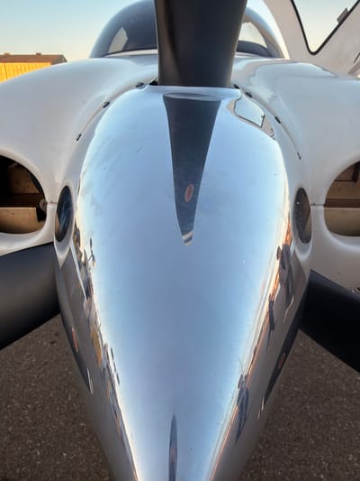 An extreme close-up, low-angle shot shows the highly reflective nose cone of a white aircraft. The nose cone is made of polished metal and reflects the surrounding environment. In the reflection, the sky is visible at the top, transitioning from a light blue to a warm orange and yellow hue near the horizon. Below the sky, a building with a wooden exterior is visible, along with parts of the aircraft's canopy. The reflection also shows the black propeller, which has a red stripe and a white stripe near the tip, and the aircraft's body. The ground, appearing as asphalt with small gravel, is visible at the bottom of the reflection. Several indistinct figures of people are also reflected in the nose cone, appearing as dark silhouettes. The scene is set outdoors, likely at an airport, given the presence of the aircraft and the runway-like ground. The overall impression is one of detail and reflection, highlighting the smooth, polished surface of the aircraft.
