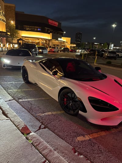 The image captures a nighttime scene at Oakbrook Center in Oak Brook, Illinois, USA. A white McLaren sports car is parked along a curb in front of Wildfire Restaurant. Adjacent to the McLaren is another white car. The foreground focuses on the McLaren's front passenger side, showcasing its sleek design, wheel, and red brake caliper. The background includes the illuminated facade of the upscale shopping center with restaurants like Wildfire and other storefronts visible. Cars are parked in the background along the street. A skyscraper is visible behind buildings. The overall lighting is a mix of the restaurant lights and streetlights.