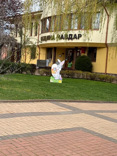 An outdoor daytime scene depicts a building, a lawn, and a paved walkway. The image is oriented with the building on the left, a strip of green grass in the middle, and a patterned brick walkway on the right.
The building, light yellow or beige in color, features multiple windows and a brown gutter system. Large, dark Cyrillic letters spelling "АВДА" are mounted on its facade, partially obscured by a leafy tree branch. Near one of the building's entrances, a small red sign with white text reads "БАНКОВ ATM" (BANKOV ATM). A person wearing a green shirt is partially visible through an open doorway or window of the building, suggesting activity inside or at the entrance.
On the manicured green lawn in front of the building, a large, colorful cardboard cutout of a grey bunny holding a basket filled with Easter eggs is prominently displayed, indicating the spring season and possibly the time around Easter. The lawn also shows a few small yellow flowers.
The paved walkway features a geometric pattern of interlocking bricks in shades of beige, reddish-brown, and dark grey. Sparse trees with budding leaves and some bare branches are scattered throughout the scene, further reinforcing the spring setting. The lighting suggests a clear day, likely late morning or early afternoon. The visible text and location context point to a public or commercial building in Chavdar, Bulgaria.