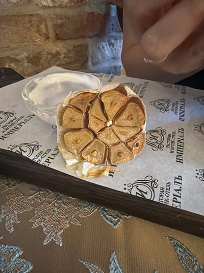 The image displays a close-up of a roasted head of garlic, cut in half, resting on a white paper placemat with a repeating pattern of Russian text that reads "РЕСТОРАН И БУТИК-ОТЕЛЬ ИМПЕРИАЛЬ". A small glass dish containing a white creamy substance, possibly a dip or sauce, is visible to the left of the garlic. A dark wooden serving board is positioned diagonally across the bottom of the frame. In the background, a textured brick wall and a person's hand with a ring on one finger are partially visible. The scene is set on a table covered with a brown cloth featuring an ornate embroidered pattern. This setting is consistent with a restaurant or hotel dining area, likely within the city of Suzdal, Russia, given the Russian text on the placemat.
