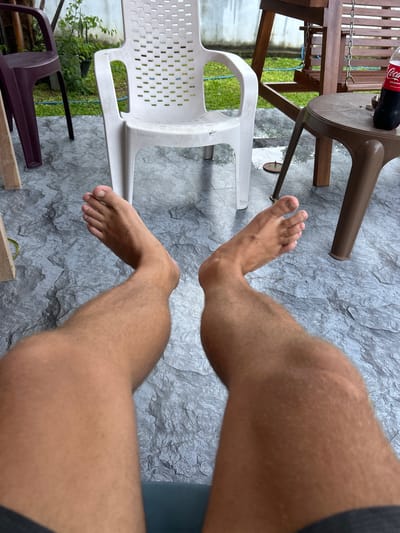 The image shows a first-person perspective of a person sitting outdoors in Kadewatta, Sri Lanka. The subject's legs and feet are visible, positioned in the foreground. The person appears to be wearing dark shorts. Their right leg is bent, with the foot resting on a white plastic chair. The left leg is also bent, with the foot positioned near a dark brown plastic chair.
The background consists of a textured grey stone-like floor. Behind the chairs, there is a wooden table with a red Coca-Cola bottle on it, and some greenery, suggesting an outdoor seating area, possibly a patio or garden. The overall lighting suggests it is daytime, with no specific weather conditions being evident. The scene is casual and relaxed, with the focus on the person's lower body and their immediate surroundings. There is no discernible text other than the label on the Coca-Cola bottle, which reads "Coca-Cola Original Taste".