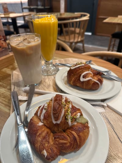 This image shows two croissants and two drinks on a wooden table in what appears to be a cafe in Oviedo/Uviéu, Spain. The croissants are on white plates, and each has white icing and chopped pistachios on top. One croissant is filled with a green cream, possibly pistachio cream. The drinks are in tall glasses. One is an iced coffee with milk, and the other is a bright orange beverage, likely juice. Cutlery, including forks and knives, are placed next to the croissants. In the background, wooden chairs and tables are visible, suggesting the setting of a cafe or restaurant. The lighting suggests it is daytime.