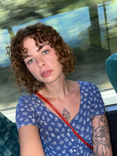A woman with curly brown hair and green eyes is pictured from the chest up, looking forward. She is wearing a blue dress with a white floral pattern and a red strap across her chest. She has a tattoo on her chest and arm. The background is blurred, suggesting movement, and appears to be a natural landscape with trees and water. The image appears to have been taken from a moving vehicle, possibly a bus or train, as indicated by the window and seating. The scene is likely Perazića Do, Montenegro, given the context provided.