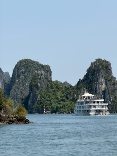 This image captures a picturesque scene of a large cruise ship navigating or anchored within a bay surrounded by dramatic limestone karsts. The main subject is a multi-decked white cruise ship, visible in the mid-ground, nestled between the towering rock formations. The ship features numerous windows, balconies, and railings, and a red flag with a yellow star, the national flag of Vietnam, is visible near its upper deck.

The bay is characterized by its distinctive karst islands, which rise steeply from the calm, blue-green water. These islands are heavily vegetated with dense green trees and shrubs, contrasting with the grey, rugged rock faces. In the mid-ground, on one of the smaller islands, a yellow diamond-shaped warning sign with a black border and a black exclamation mark is clearly visible, likely indicating a hazard or caution area for navigation.

The water surface is relatively calm, showing subtle ripples, reflecting the clear, pale blue sky that dominates the upper portion of the image. The lighting suggests a bright, clear day, likely late morning or early afternoon, with no harsh shadows. The overall setting evokes a sense of tranquility and natural grandeur, typical of the iconic landscapes found in areas such as Ha Long Bay in Vietnam. The scene highlights the interplay between natural beauty and human presence, with the cruise ship offering a way to experience the stunning environment.