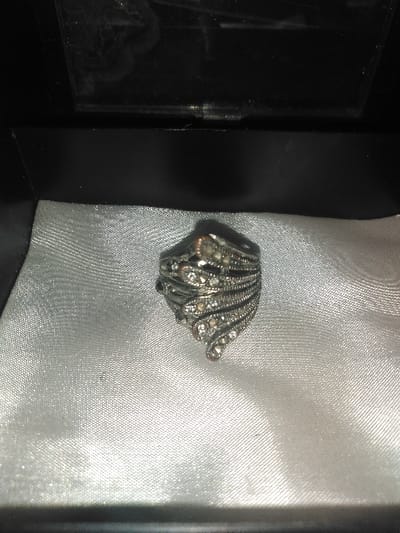 A single decorative ring is centered within an open black jewelry box in Gerasimovskoye, Kazakhstan. The ring is crafted from silver-toned metal, featuring multiple intertwining, wave-like bands adorned with numerous small, clear, faceted stones. The interior of the jewelry box is lined with a lightly textured, white or silver-white satin-like fabric. The inner surface of the box lid, visible at the top of the frame, is black and reflective, showing minor smudges or dust. No individuals, actions, or interactions are present in the scene.