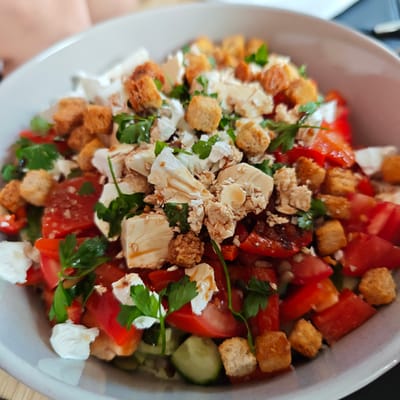 A close-up, eye-level shot shows a bowl of salad. The salad contains cubed white cheese, chopped red tomatoes, sliced cucumber, and croutons. Fresh green parsley is scattered throughout the salad, and a light dressing is drizzled over the top. The salad is presented in a light grey bowl. The background is blurred, showing a hint of a person's arm and shoulder in the upper left, and a dark surface in the upper right. The scene is indoors, likely a dining setting. There are no identifying landmarks or features to suggest a specific location within Locking Parklands, United Kingdom.