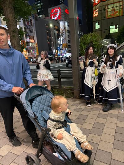 In Chiyoda, Japan, at night, a man stands to the left, pushing a stroller with a toddler inside. The man wears a blue jacket and black pants. The toddler is dressed in a striped shirt and light-colored pants, wearing sandals. Behind them, two young women in maid uniforms stand on a sidewalk. The woman on the left has purple-tinted hair and holds a book. The woman on the right has dark hair and wears a maid headdress. Both women are smiling. The background features illuminated buildings with bright signs, typical of a busy urban environment. A large tree is visible to the left of the man. The ground is paved with interlocking bricks.