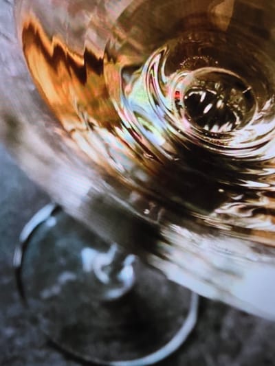 The image shows a close-up of a glass with a liquid, likely alcoholic, swirling in it. The lighting creates reflections and distortions, giving the liquid an iridescent quality. The background is dark and textured, possibly a table or countertop. The location in the city of Voluntari, Romania, is not discernible from the image itself. There are no people or discernible actions or interactions visible. The scene is abstract and focuses solely on the visual properties of the liquid and the glass.