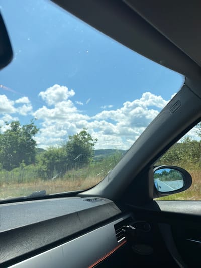 The media file is a photograph taken from inside a car, looking out through the windshield and side window. The perspective is tilted, suggesting the car might be on an incline or the photo has been rotated. The scene outside the car is a rural landscape with a bright blue sky filled with fluffy white clouds. Trees and vegetation are visible, suggesting a natural, possibly wooded or grassy area. In the foreground, inside the car, a portion of the dashboard is visible, including a side mirror and what appears to be an air vent. The side mirror reflects a bit of the sky and some greenery. The interior of the car appears to be dark, with some textured surfaces. There is visible text on the car's interior pillar that reads "AIRBAG". The image appears to be taken during the daytime, likely on a clear and sunny day. The overall impression is one of a journey through a scenic, rural environment.