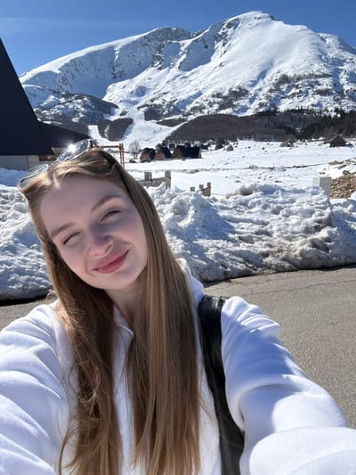 The image presents a young adult female captured in a selfie in a snowy, mountainous landscape identified as Virak, Montenegro. She occupies the foreground, looking directly at the camera with her left eye winking and a slight smile. Her long, light brown hair cascades down, and she wears a white zippered hooded top. Sunglasses are perched on top of her head. A black strap, likely from a bag, is visible over her left shoulder.

The midground features a grey asphalt road flanked by irregular piles of melting, dirty snow. Beyond the snow, low stone walls and scattered small wooden cabins or chalets with dark roofs are visible across the snow-covered terrain. In the background, large, prominent snow-capped mountains dominate the horizon under a clear blue sky. A ski slope is discernible on one of the mountain faces, alongside areas of sparse deciduous trees and evergreen foliage. A section of a large, dark-roofed building with a steep pitch is visible on the far left of the frame. The scene is brightly illuminated by direct sunlight.