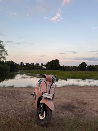 A light pink scooter is parked on a dirt path next to a body of water with lush green vegetation and trees in the background. The sky is a pale blue with streaks of pink and white clouds, suggesting it is either early morning or late evening. The scooter has a license plate that reads "BL 6790 KBH". The scene appears to be in a rural or natural setting, possibly Matangsijuek Teungoh, Indonesia, given the vegetation and the style of the license plate.