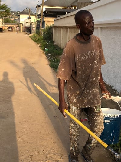 A man stands outdoors, coated in white paint splatters on his brown t-shirt and pants. He holds a yellow pole, appearing to be a painting tool. Beside him is a blue and white bucket. The setting is a street with buildings visible in the background, suggesting an urban or residential area. The angle and shadow indicate it's daytime, likely in the late afternoon. The scene conveys a sense of work and activity, with the man possibly engaged in painting a building or structure nearby.