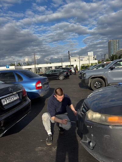 A man is working on a dark-colored car bumper in a parking lot. He is kneeling on the asphalt, wearing a dark blue long-sleeved shirt and grey sweatpants, with white socks and black sneakers. He holds a screwdriver and is working on the front bumper of the car.

Several other cars are parked in the lot, including a grey SUV and a blue hatchback. In the background, there are buildings and a cloudy sky. The image appears to have been taken during the daytime.

The license plate on the black car in the foreground reads "P910XX 797 RUS". There is also a blue parking sign visible. The overall scene suggests a casual repair or adjustment being made to a vehicle in an outdoor parking area.