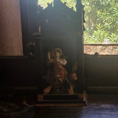 The image shows a small, dark wooden shrine with a figure of a saint or angel inside. The figure is dressed in red and white robes and has a halo. The shrine is placed on a dark surface, possibly a table or shelf. In the background, there is a window with a view of green trees. The lighting is dim, with a bright light coming from the window. The shrine appears to be old and possibly religious in nature. It is difficult to determine the specific location within Lloret de Mar, Spain, without additional context or identifying landmarks.