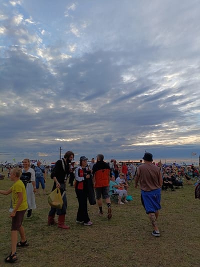 The image depicts a gathering of people outdoors, under a dramatic, cloudy sky. The setting appears to be a festival or an event in Lagunovo, Russia. In the foreground, a young boy in a yellow t-shirt and dark shorts is walking to the left. Behind him, a woman in a white dress and a man in a black shirt and red boots are conversing. Further back, a group of people are milling around, with tents and flags visible in the distance, suggesting a fairground or festival site. The overall atmosphere is casual and lively. The ground is covered in dry grass. The lighting indicates it is likely late afternoon or early evening, with the sun's rays breaking through the clouds.