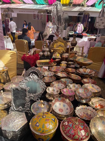 The image shows a variety of decorative brass and silver items on display at a market stall in Chandigarh, India. The stall is located at the coordinates 30.764° N, 76.798° E. The stall is set up outdoors under a canopy of colorful fabric. The items on display include bowls, plates, trays, mirrors, boxes, and other decorative pieces. The bowls are decorated with intricate floral designs in vibrant colors. The mirrors are ornately framed in silver.  There is an assortment of small decorative boxes. The scene is bustling with activity as people browse the items on display. The stall is located near a bustling street in Chandigarh, India.  The stall is covered by a white canopy with a red, yellow, and blue awning.  There are other stalls and booths in the background. Several people are visible in the image. People are looking at the items, buying items, and talking to the vendors. There is a large, industrial-style fan in the background. The stall is located in the midst of a busy market in Chandigarh, India.