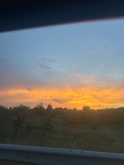 The image shows a sunset over a rural landscape. The sky is a gradient of blue at the top, transitioning to bright orange and yellow as it meets the horizon. Clouds are visible, illuminated by the setting sun. Below the sky is a silhouette of trees and vegetation. In the foreground, a dark railing runs horizontally across the bottom of the frame. The scene appears to be captured from a moving vehicle, possibly on a road. The location is identified as Zagorsko, Bulgaria.