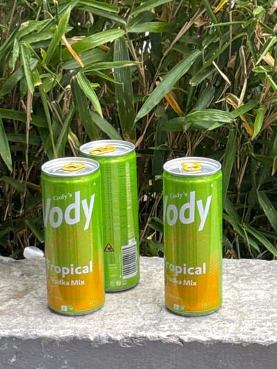 The image shows three discarded cans of "Cody's Tropical Vodka Mix" resting on a gray concrete surface. The cans are partially surrounded by green leafy vegetation, resembling bamboo or similar plant life. The scene appears to be outdoors. The information about the location of the city of Orsay, France is not available in the picture.