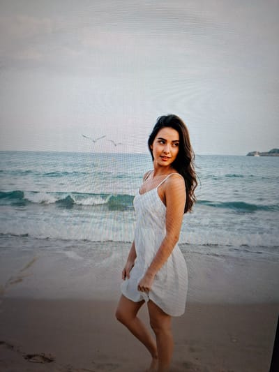 A young woman with long, dark hair stands on a sandy beach, looking over her shoulder towards the right of the frame. She is wearing a white, spaghetti-strap dress. The ocean is behind her, with waves gently rolling onto the shore. The sky is overcast and the lighting suggests it is either early morning or late evening. There are some faint details of distant land or structures on the horizon. There is no indication of the city of Tenevo, Bulgaria in this image.