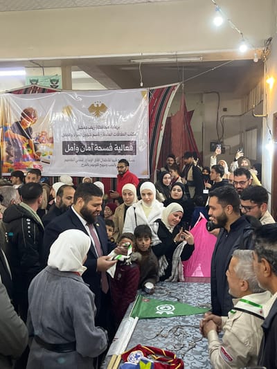 An indoor scene in Kafr Batna, Syria, depicts a crowded community event. The room has light-colored walls, fluorescent lighting, and string lights overhead. A large white banner on the back wall features Arabic text, including "برعاية محافظة ريف دمشق" (Under the patronage of Damascus Countryside Governorate) and "فعالية فسحة أمان وأمل" (Activity: Space for Safety and Hope), alongside a graphic of a child playing with blocks and a golden eagle emblem (Syrian coat of arms). Flanking the banner are patterned fabric drapes or tent-like structures.
In the immediate foreground, a man in a dark suit with a beard is handing a small white and green toy airplane to a woman wearing a grey coat and a white hijab. A young child with faint face paint resembling whiskers stands beside them, observing the interaction. To the right, another man in a dark jacket is shaking hands with an older man dressed in a khaki shirt with a green scarf and badges, typical of a scout uniform.
A table covered with a patterned cloth runs along the foreground, displaying various objects including small green flags with a white star, crescent, and ribbon emblem, and multiple small metal items resembling clasps or hooks. Several scout-style neckerchiefs and badges are also visible on the table.
The background is densely populated with men, women, and children. Many women are wearing hijabs. Individuals are observing the activities, conversing, and some are holding up smartphones to record or photograph the event. The overall atmosphere appears organized and engaged.