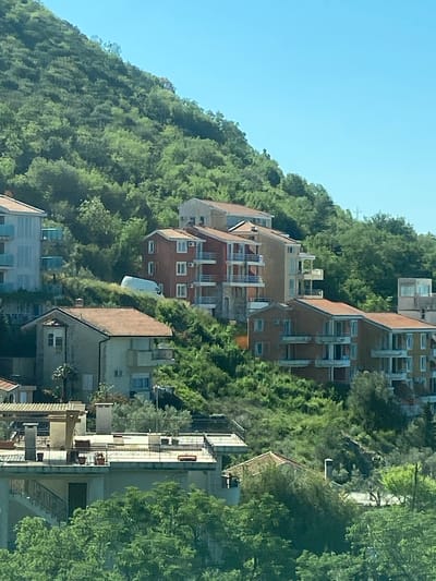 The image depicts a hillside neighborhood in Prijevorac, Montenegro. Terraced residential buildings are nestled amongst dense vegetation. The buildings range in size and architectural style, with several multi-story structures featuring red brick facades and balconies. Lush greenery covers the hillside, partially obscuring some of the buildings. A clear, blue sky is visible in the upper left corner of the image.