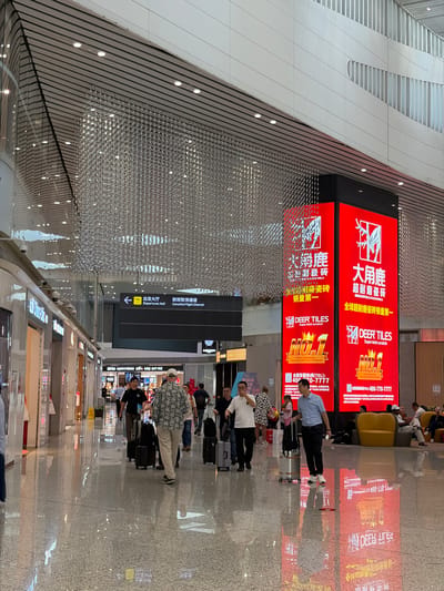 The image depicts the interior of Guangzhou Baiyun International Airport in Baiyun District, China. A large red advertisement for "Deer Tiles" is prominently displayed, showing both Chinese and English text and contact information. People are seen waiting with luggage, likely passengers. A sign indicates "Departures Hall" and "Cancelled Flight Channel." Shops are visible in the background. The ceiling has a complex, modern design.